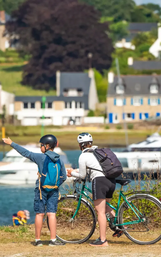 De foto toont twee gehelmde mensen met fietsen, op de voorgrond bij het water, met boten en huizen op de achtergrond.