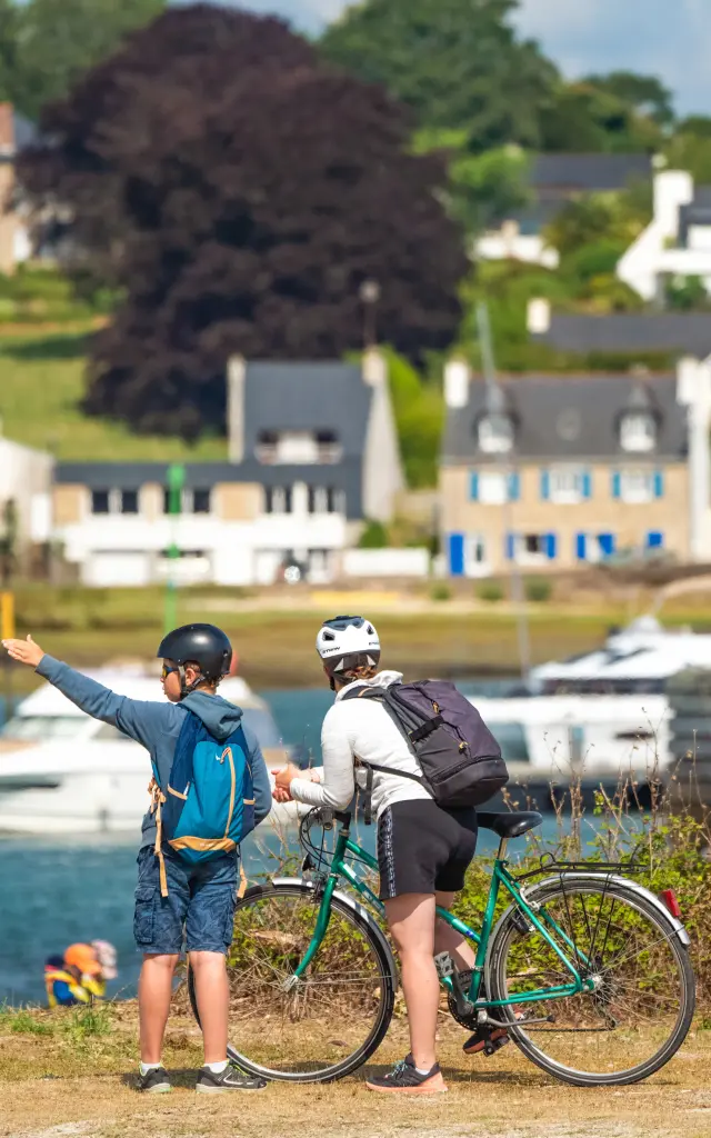 La photo montre deux personnes casquées avec des vélos, au premier plan près de l’eau, avec des bateaux et des maisons en arrière-plan.
