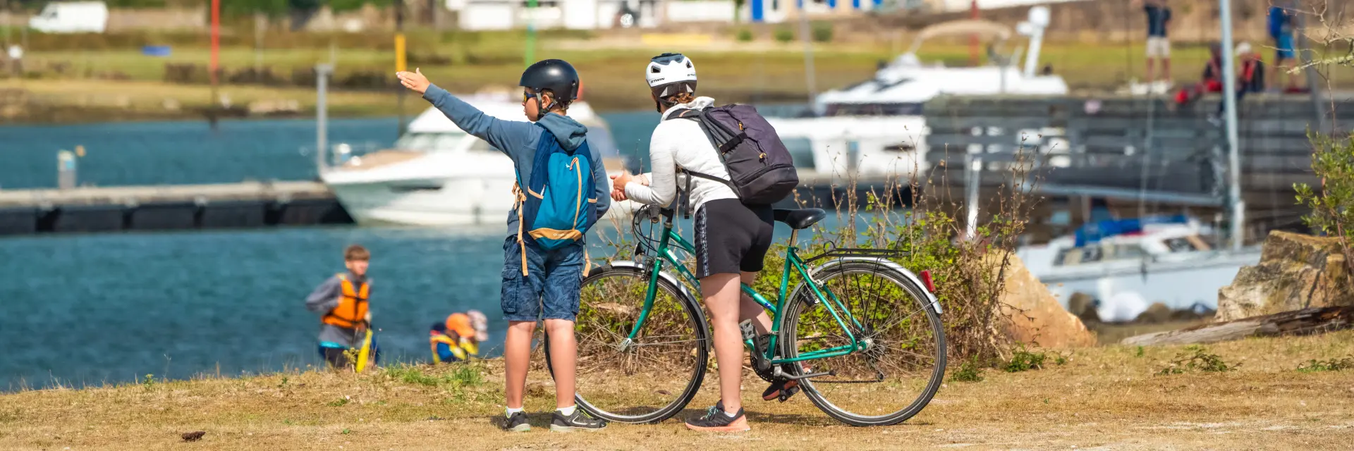 La photo montre deux personnes casquées avec des vélos, au premier plan près de l’eau, avec des bateaux et des maisons en arrière-plan.
