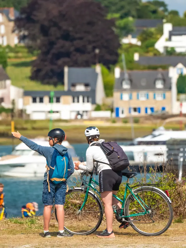 La photo montre deux personnes casquées avec des vélos, au premier plan près de l’eau, avec des bateaux et des maisons en arrière-plan.