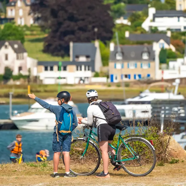 La photo montre deux personnes casquées avec des vélos, au premier plan près de l’eau, avec des bateaux et des maisons en arrière-plan.