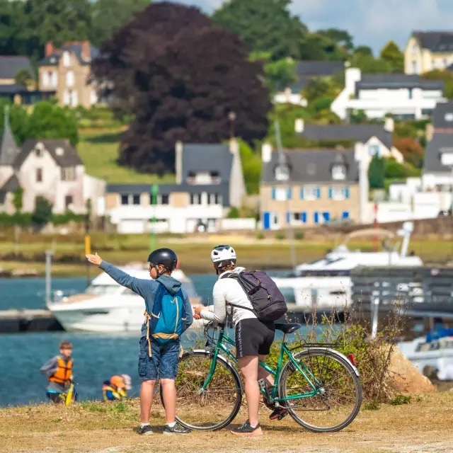 La photo montre deux personnes casquées avec des vélos, au premier plan près de l’eau, avec des bateaux et des maisons en arrière-plan.