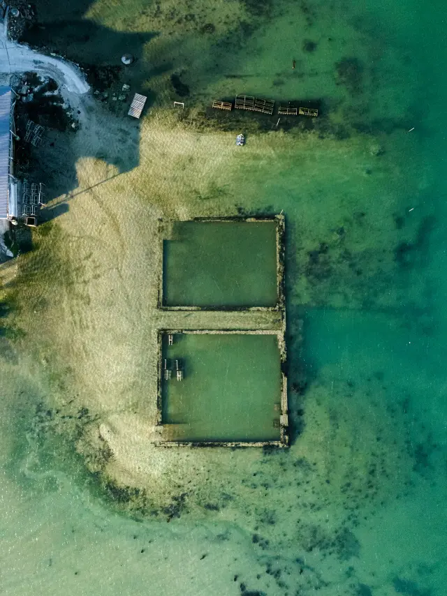 Une photo aérienne qui montre une zone côtière avec eau turquoise/verte et des bancs de sable. Deux bassins rectangulaires (type viviers) sont visibles au centre-bas de l’image, entourés de structures sombres. En haut à gauche de la photo, une zone de rivage et quelques éléments alignés : bâtiments et caisses apparaissent.