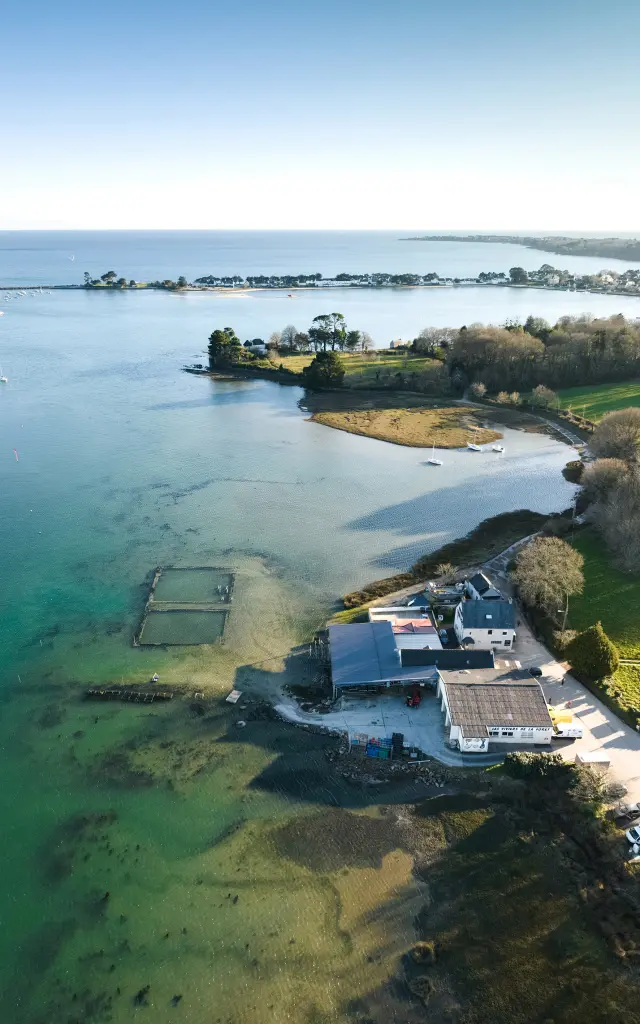 Une photo qui montre une vue aérienne d’une côte: mer bleu-vert au premier plan, zones peu profondes visibles sous l’eau, un petit bâtiment au bord de l’eau avec des viviers, une bande de terre avec des arbres, puis la mer et le littoral au loin sous un ciel bleu.