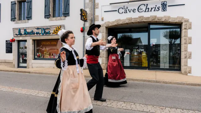Ein Foto, das drei Personen in traditionellen bretonischen Trachten (Kleider und Westen) zeigt, die auf einer Straße vor Geschäften tanzen. Auf einem Schild über einem großen Schaufenster steht La Cave de Chris. Ein weiteres Geschäft auf der linken Seite hat ein Schild mit der Aufschrift La Boucle en Vrac und im Schaufenster steht Épicerie vrac.