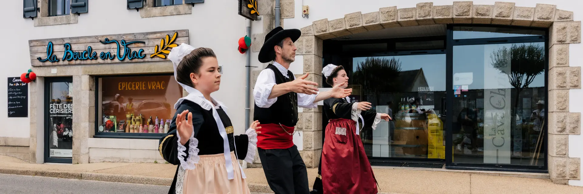 Une photo qui montre trois personnes en costumes traditionnels bretons (robes et gilets) en train de danser dans une rue, devant des commerces. Une enseigne au-dessus d’une grande vitrine indique “La Cave de Chris”. Un autre commerce, à gauche, a une enseigne “La Boucle en Vrac”, et la vitrine porte “Épicerie vrac”.