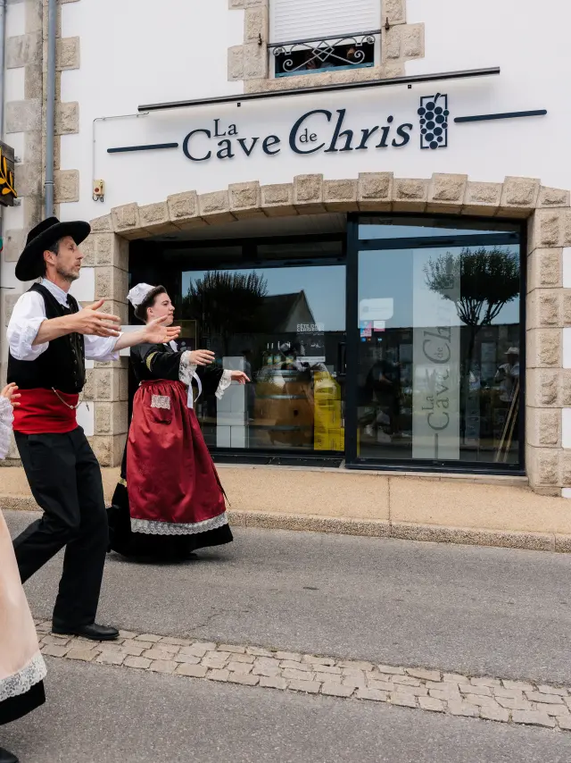 Une photo qui montre trois personnes en costumes traditionnels bretons (robes et gilets) en train de danser dans une rue, devant des commerces. Une enseigne au-dessus d’une grande vitrine indique “La Cave de Chris”. Un autre commerce, à gauche, a une enseigne “La Boucle en Vrac”, et la vitrine porte “Épicerie vrac”.