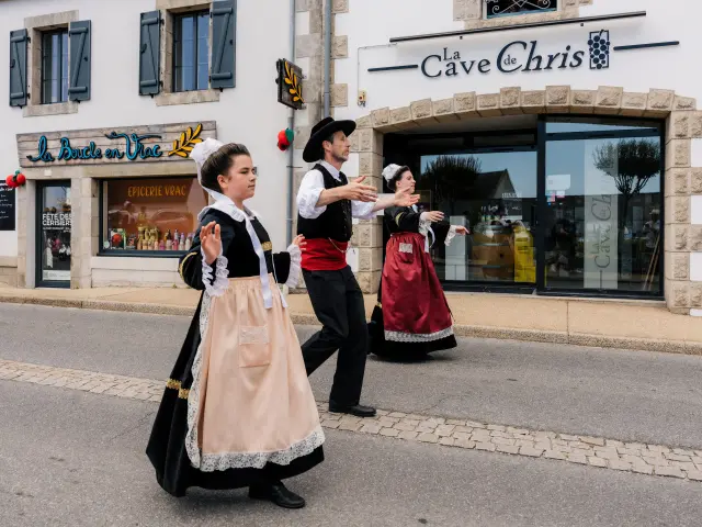 Une photo qui montre trois personnes en costumes traditionnels bretons (robes et gilets) en train de danser dans une rue, devant des commerces. Une enseigne au-dessus d’une grande vitrine indique “La Cave de Chris”. Un autre commerce, à gauche, a une enseigne “La Boucle en Vrac”, et la vitrine porte “Épicerie vrac”.