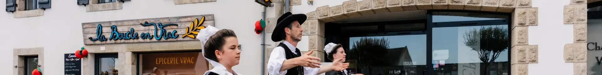 Een foto waarop drie mensen in traditionele Bretonse klederdracht (jurken en vesten) dansen in een straat voor winkels. Op een bord boven een grote etalage staat La Cave de Chris. Een andere winkel, aan de linkerkant, heeft een bordje La Boucle en Vrac en op het raam staat Épicerie vrac.