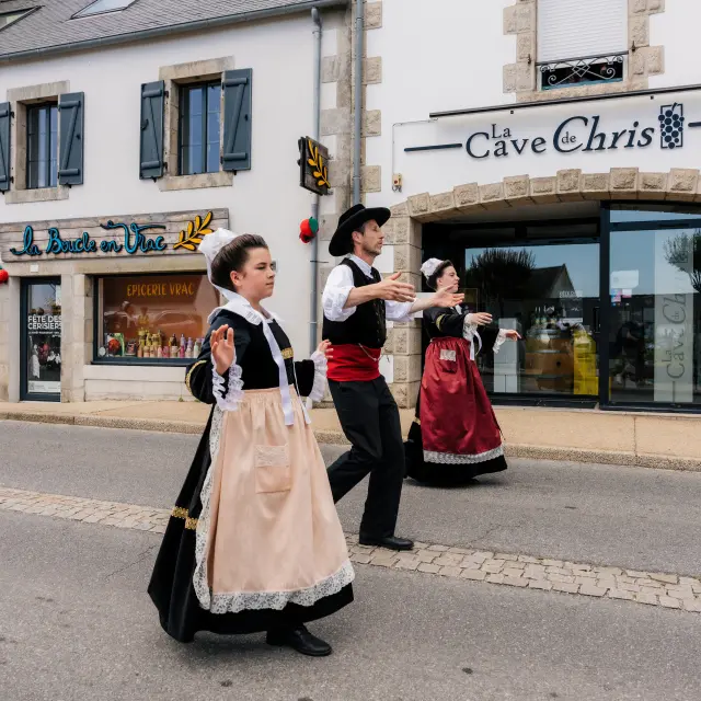 Une photo qui montre trois personnes en costumes traditionnels bretons (robes et gilets) en train de danser dans une rue, devant des commerces. Une enseigne au-dessus d’une grande vitrine indique “La Cave de Chris”. Un autre commerce, à gauche, a une enseigne “La Boucle en Vrac”, et la vitrine porte “Épicerie vrac”.