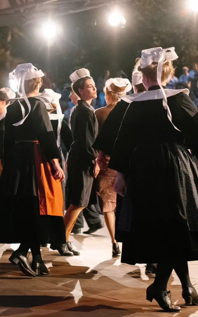 Une photo qui montre un groupe d’adultes en tenue traditionnelle bretonne sombre (robes et tabliers pour plusieurs femmes, gilets/chemises pour certains hommes) en train de danser sous une structure couverte, éclairée par plusieurs projecteurs. Plusieurs danseuses portent une coiffe blanche avec rubans. Un public est visible en arrière-plan dans l’obscurité, avec des lumières chaudes.