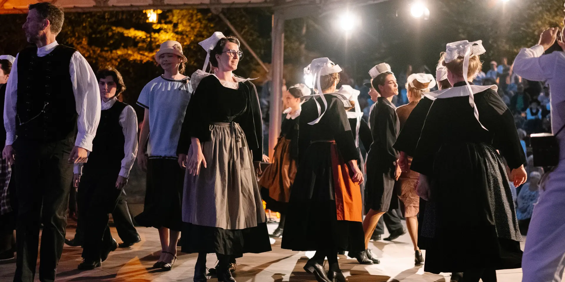 A photo showing a group of adults in dark Breton traditional dress (dresses and aprons for several women, waistcoats/shirts for some men) dancing under a covered structure lit by several spotlights. Several of the dancers are wearing white headdresses with ribbons. An audience is visible in the dark background, with warm lights.