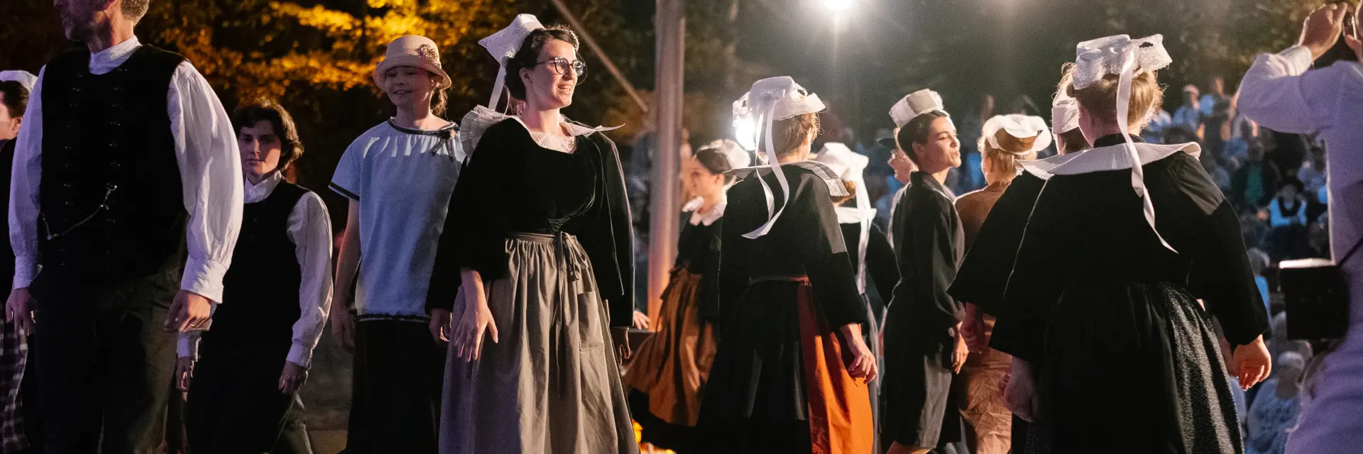 Une photo qui montre un groupe d’adultes en tenue traditionnelle bretonne sombre (robes et tabliers pour plusieurs femmes, gilets/chemises pour certains hommes) en train de danser sous une structure couverte, éclairée par plusieurs projecteurs. Plusieurs danseuses portent une coiffe blanche avec rubans. Un public est visible en arrière-plan dans l’obscurité, avec des lumières chaudes.