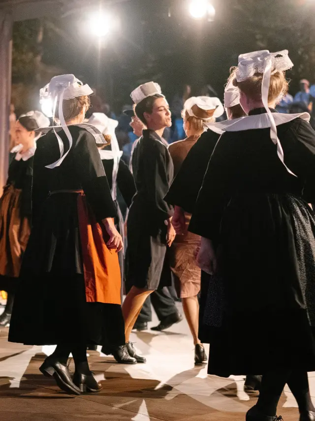 A photo showing a group of adults in dark Breton traditional dress (dresses and aprons for several women, waistcoats/shirts for some men) dancing under a covered structure lit by several spotlights. Several of the dancers are wearing white headdresses with ribbons. An audience is visible in the dark background, with warm lights.
