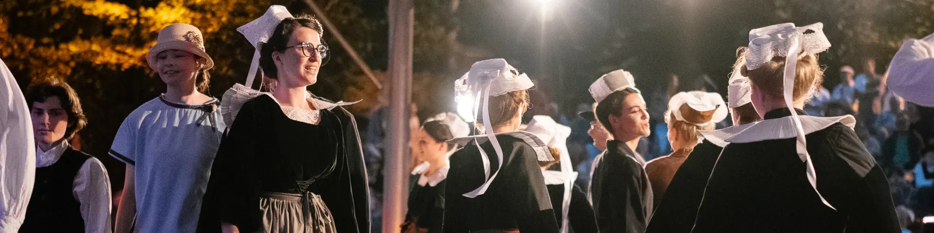 A photo showing a group of adults in dark Breton traditional dress (dresses and aprons for several women, waistcoats/shirts for some men) dancing under a covered structure lit by several spotlights. Several of the dancers are wearing white headdresses with ribbons. An audience is visible in the dark background, with warm lights.