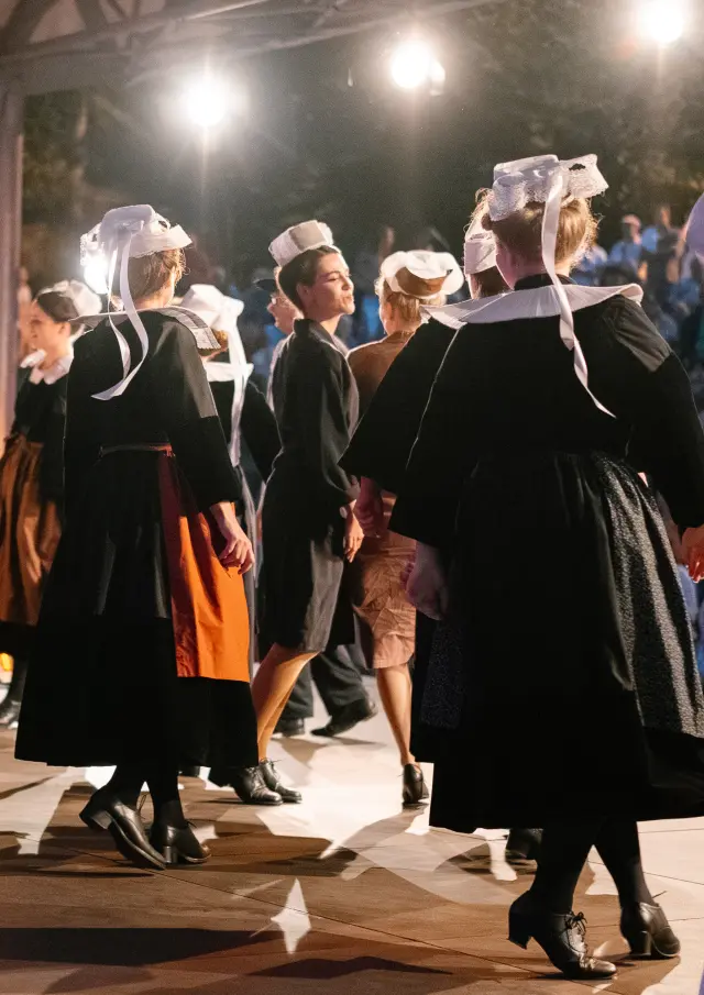 Ein Foto, das eine Gruppe von Erwachsenen in traditioneller, dunkler bretonischer Kleidung (Kleider und Schürzen bei mehreren Frauen, Westen/Hemden bei einigen Männern) zeigt, die unter einer überdachten Struktur, die von mehreren Scheinwerfern beleuchtet wird, tanzen. Mehrere Tänzerinnen tragen einen weißen Kopfschmuck mit Bändern. Im Hintergrund ist in der Dunkelheit ein Publikum zu sehen, das mit warmen Lichtern beleuchtet wird.