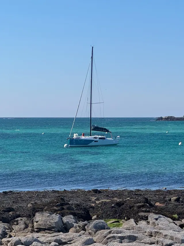 Une photo de mer qui montre un voilier à l’ancre sur une eau turquoise, sous un ciel bleu, avec une côte rocheuse au premier plan et de petites îles à l’horizon.