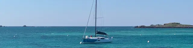 Een foto van een zeilboot voor anker in turquoise water, onder een blauwe lucht, met een rotsachtige kustlijn op de voorgrond en kleine eilandjes aan de horizon.