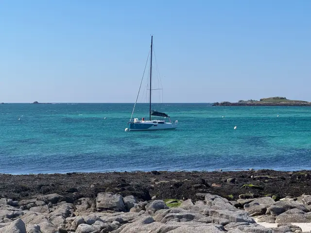 Une photo de mer qui montre un voilier à l’ancre sur une eau turquoise, sous un ciel bleu, avec une côte rocheuse au premier plan et de petites îles à l’horizon.
