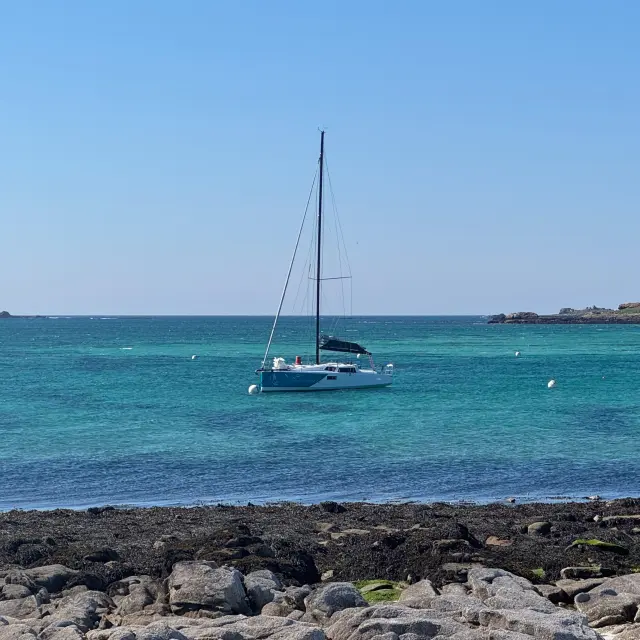 Ein Meeresfoto, das ein ankerndes Segelboot auf türkisfarbenem Wasser unter blauem Himmel zeigt, mit einer felsigen Küste im Vordergrund und kleinen Inseln am Horizont.
