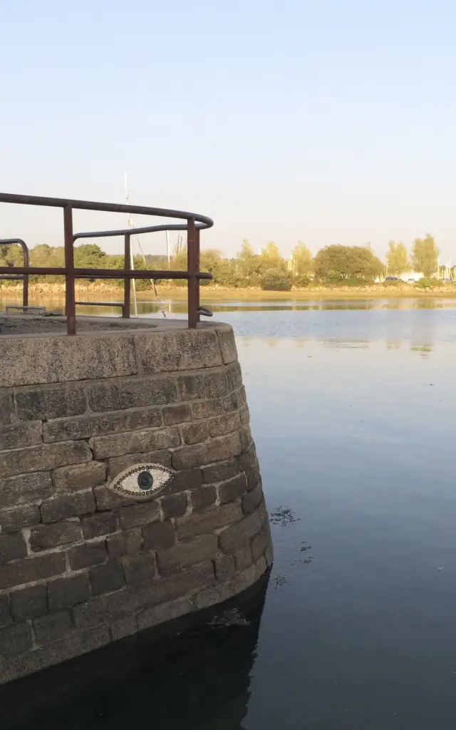 Das Foto zeigt eine Szene am Wasser. Im Vordergrund links befindet sich ein abgerundeter Steinkai mit einem Metallgeländer am oberen Ende. An der Steinmauer ist ein Augenmotiv angebracht. Zwei Personen sitzen auf einer Bank auf dem Steg. Das Wasser ist ruhig und nimmt den Großteil des Bildes auf der rechten Seite ein. Im Hintergrund ist ein Ufer mit Bäumen und weiter hinten ein Hafen mit Schiffsmasten unter einem klaren Himmel zu sehen.