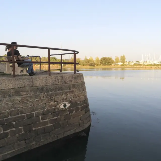 La photo montre une scène au bord de l’eau. Au premier plan à gauche, un quai en pierre arrondi avec une rambarde métallique en haut. Un motif d’œil est fixé sur le mur de pierre. Deux personnes sont assises sur un banc sur la jetée. L’eau est calme et occupe la majorité de l’image à droite. Au fond, une rive avec des arbres et, plus loin, un port avec des mâts de bateaux sous un ciel clair.