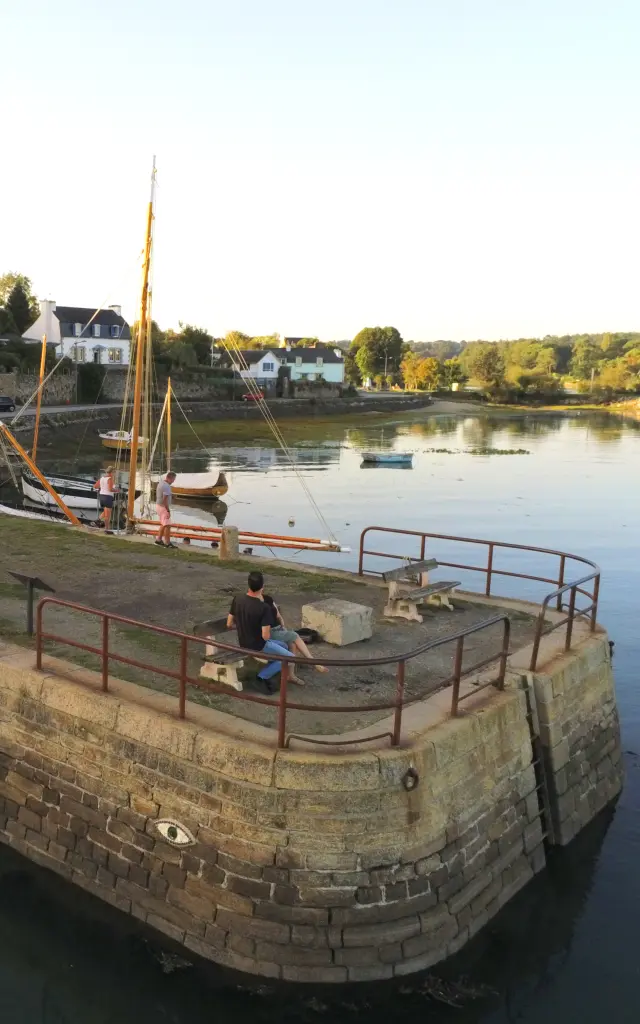 Das Foto zeigt eine Szene am Wasser. Im Vordergrund links befindet sich ein abgerundeter Steinkai mit einem Metallgeländer am oberen Ende. An der Steinmauer ist ein Augenmotiv angebracht. Zwei Personen sitzen auf einer Bank auf dem Steg. Das Wasser ist ruhig und nimmt den Großteil des Bildes auf der rechten Seite ein. Im Hintergrund ist ein Ufer mit Bäumen zu sehen, und weiter hinten sind Schiffsmasten unter einem klaren Himmel zu sehen.