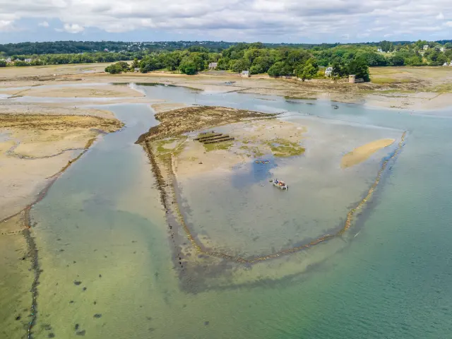 Een luchtopname van een baai bij eb met zandbanken, watergeulen, gebieden met zeewier, een kleine boot in het midden, de kust en huizen in de verte onder een bewolkte lucht.