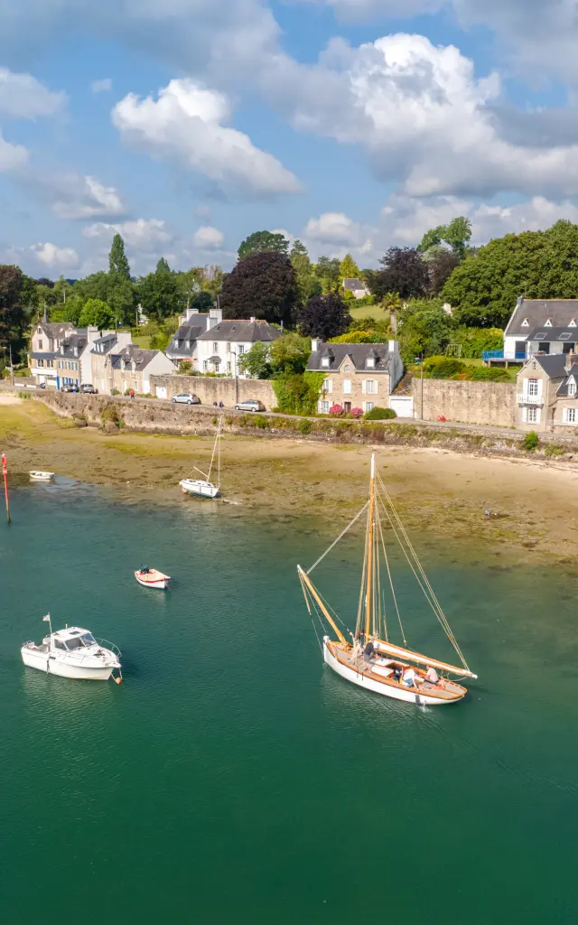 Une photo de paysage côtier qui montre la mer avec une eau vert-bleu, une bande de sable et des zones découvertes à marée basse. Plusieurs petits bateaux sont au mouillage, dont un voilier au centre et un petit bateau blanc à gauche. En arrière-plan, une rangée de maisons claires au bord de l’eau et beaucoup d’arbres verts sous un ciel bleu avec des nuages blancs.