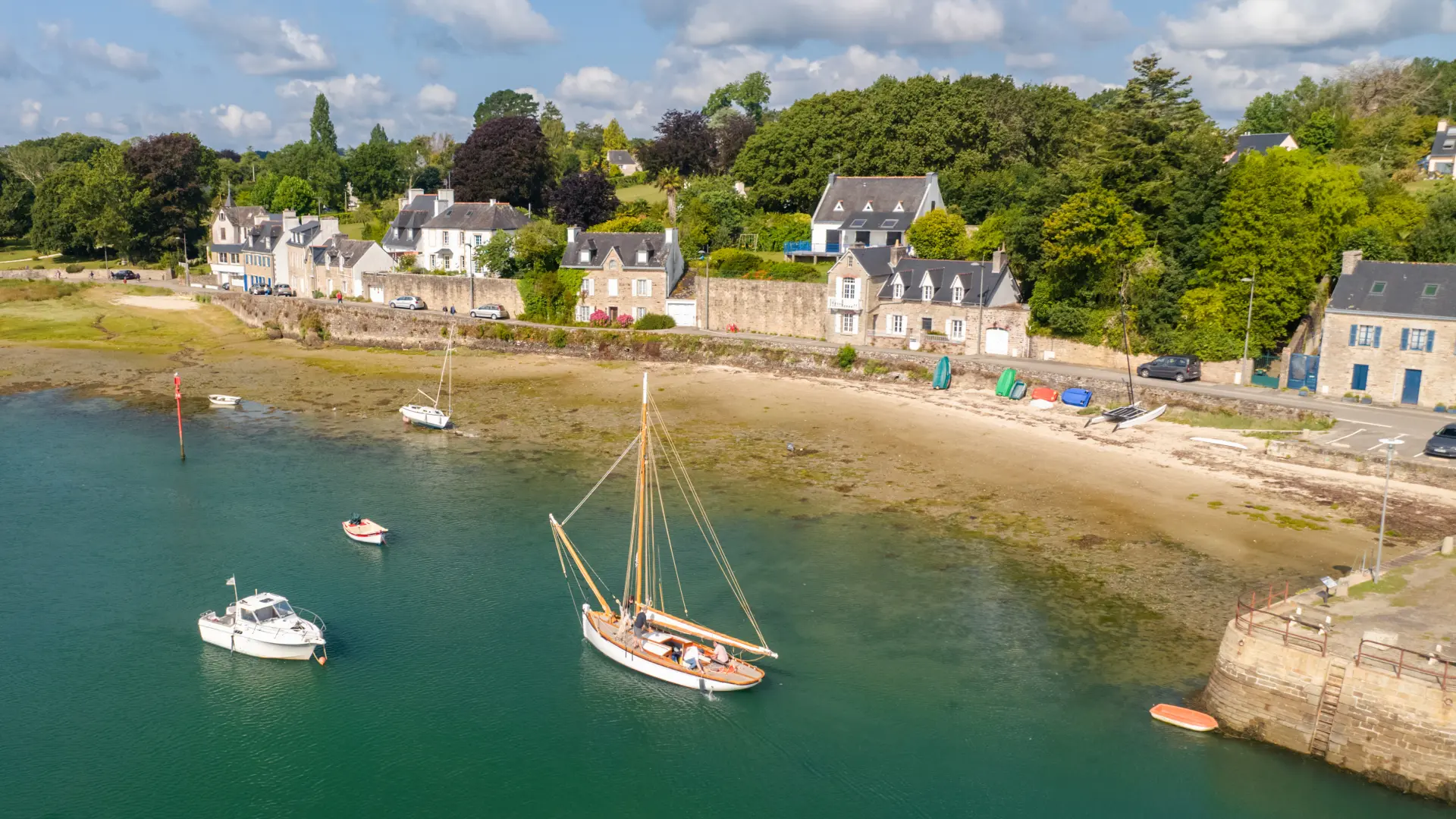 Une photo de paysage côtier qui montre la mer avec une eau vert-bleu, une bande de sable et des zones découvertes à marée basse. Plusieurs petits bateaux sont au mouillage, dont un voilier au centre et un petit bateau blanc à gauche. En arrière-plan, une rangée de maisons claires au bord de l’eau et beaucoup d’arbres verts sous un ciel bleu avec des nuages blancs.