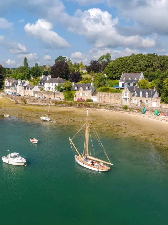 A coastal landscape shot showing the sea with green-blue water, a strip of sand and open areas at low tide. Several small boats are at anchor, including a sailing boat in the centre and a small white boat on the left. In the background, a row of light-coloured houses on the water's edge and lots of green trees under a blue sky with white clouds.