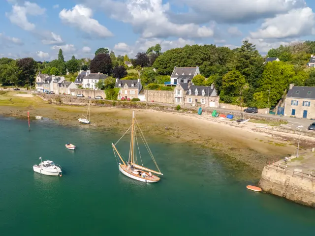 Une photo de paysage côtier qui montre la mer avec une eau vert-bleu, une bande de sable et des zones découvertes à marée basse. Plusieurs petits bateaux sont au mouillage, dont un voilier au centre et un petit bateau blanc à gauche. En arrière-plan, une rangée de maisons claires au bord de l’eau et beaucoup d’arbres verts sous un ciel bleu avec des nuages blancs.