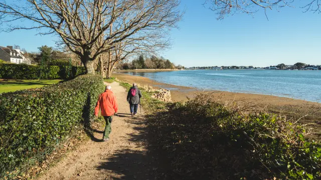 Een foto van een voetpad aan zee in de winter. Twee mensen lopen over het pad (de ene in een rode jas, de andere in een donkere jas met een roze rugzak). Links van het pad een heg en hoge, bladerloze bomen. Rechts een strand en een rustig stuk water onder een blauwe hemel.