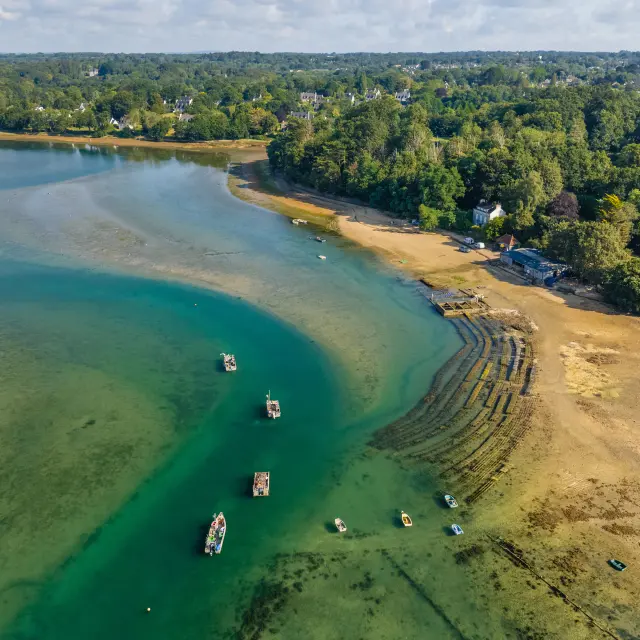 Une photo montrant une vue aérienne d’une baie à marée basse: eau turquoise et verte, grandes zones de sable découvertes, plusieurs petites embarcations sur l’eau, et au bord une zone boisée avec quelques bâtiments. Des alignements en arcs de structures sur l’estran, type parc à coquillages sont visibles près du rivage.