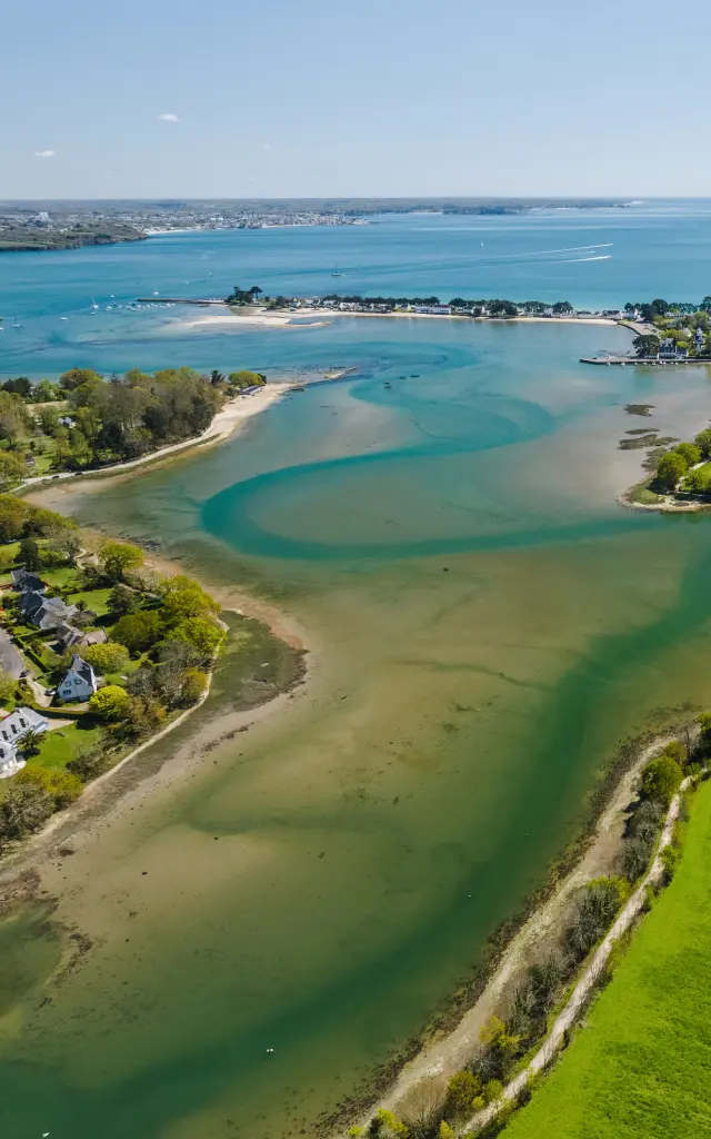 an aerial photograph in landscape. The image shows a coastal cove with sandbanks and blue-green waters, a seaside village in the centre, and large green fields in the foreground on the right; the ocean stretches to the horizon under a blue sky.