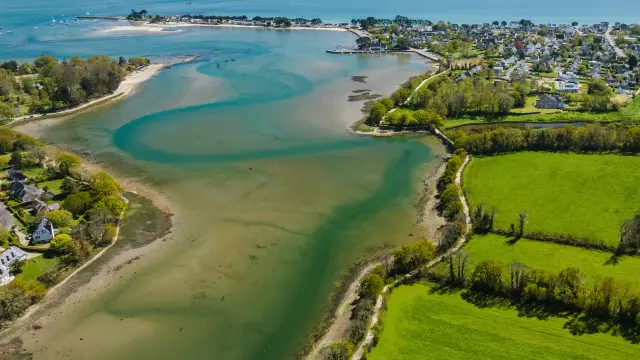 ein Luftbild als Landschaft. Das Bild zeigt eine Küstenbucht mit Sandbänken und blaugrünem Wasser, ein Dorf am Meer in der Mitte und große grüne Felder im Vordergrund rechts; der Ozean erstreckt sich am Horizont unter einem blauen Himmel.