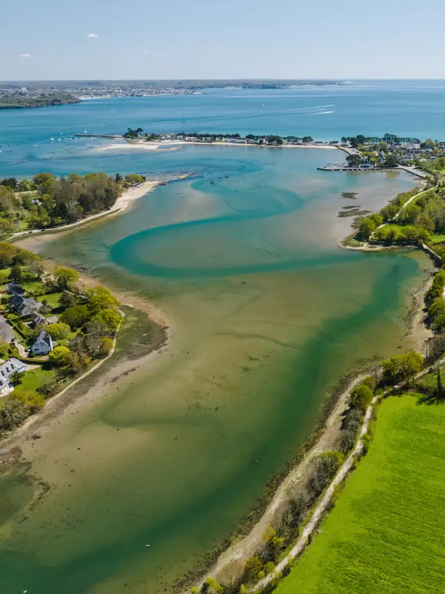 une photo aérienne en paysage. L’image montre une anse côtière avec des bancs de sable et des eaux bleu-vert, un village en bord de mer au centre, et de grands champs verts au premier plan à droite; l’océan s’étend à l’horizon sous un ciel bleu.