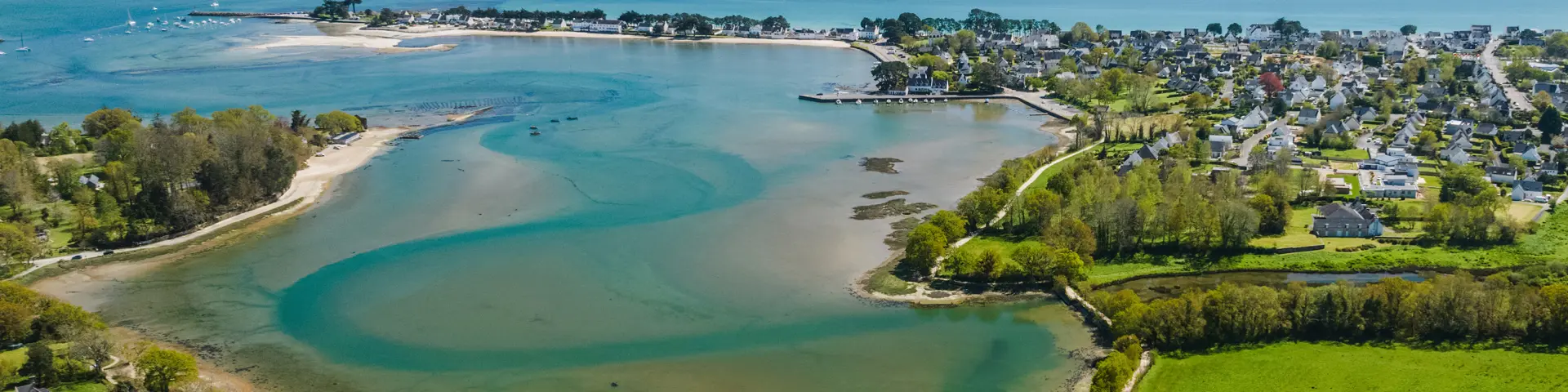 een luchtfoto in landschap. De afbeelding toont een baai aan de kust met zandbanken en blauwgroen water, een dorpje aan zee in het midden en grote groene velden op de voorgrond rechts; de oceaan strekt zich uit tot aan de horizon onder een blauwe lucht.