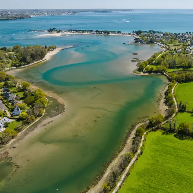an aerial photograph in landscape. The image shows a coastal cove with sandbanks and blue-green waters, a seaside village in the centre, and large green fields in the foreground on the right; the ocean stretches to the horizon under a blue sky.