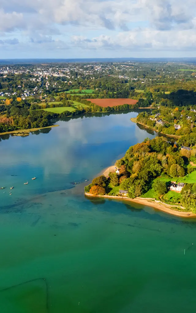 une grande photo aérienne d’une anse/côte verdoyante et d’une eau bleu-vert, sous un ciel bleu avec de gros nuages.