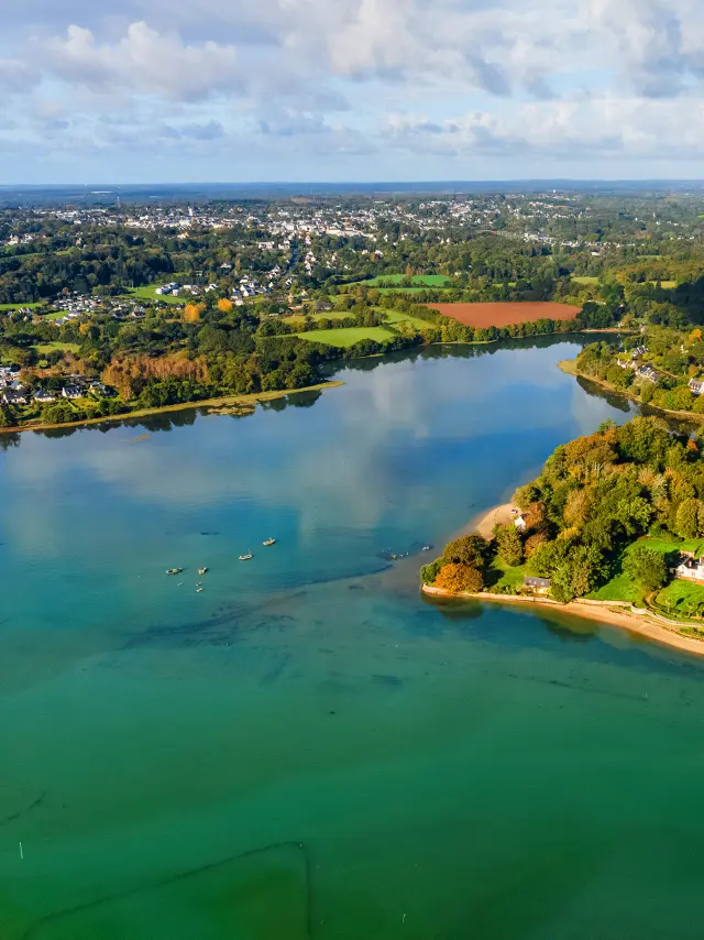 une grande photo aérienne d’une anse/côte verdoyante et d’une eau bleu-vert, sous un ciel bleu avec de gros nuages.