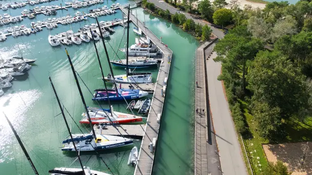 une photo aérienne au centre-gauche. La photo montre un port de plaisance rempli de bateaux à gauche, une digue/route au milieu, une zone boisée à droite, et un grand plan d’eau à droite avec une plage courbe; au fond, des maisons et de la végétation sous un ciel nuageux.
