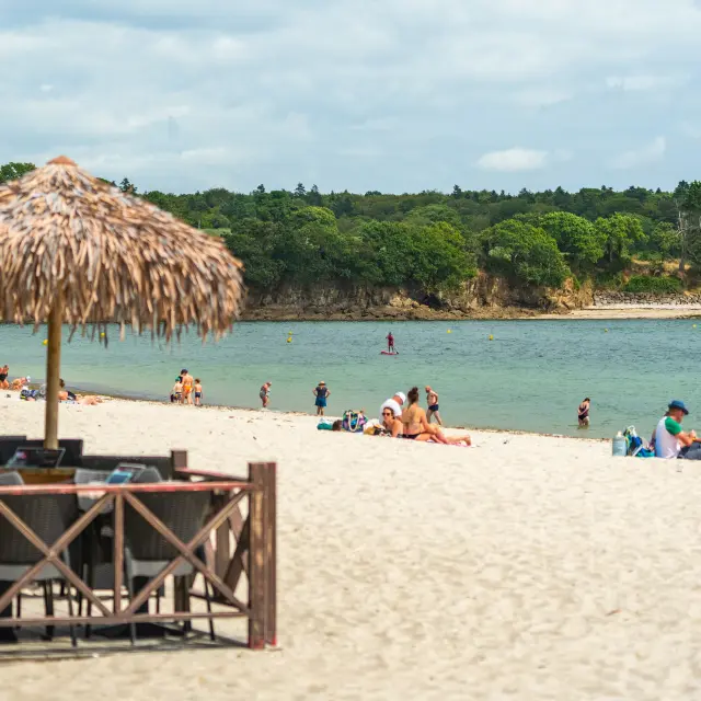 The photo shows a sandy beach with several people. On the left, a wooden bar/restaurant terrace with tables and chairs, and straw parasols. The sea is calm, and in the background a wooded coastline under a cloudy sky.