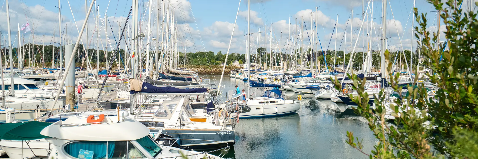 une photo au centre-gauche montrant un port rempli de voiliers et de bateaux à moteur, beaucoup de mâts, de l’eau calme, un ciel bleu avec des nuages, et de la végétation verte au premier plan.
