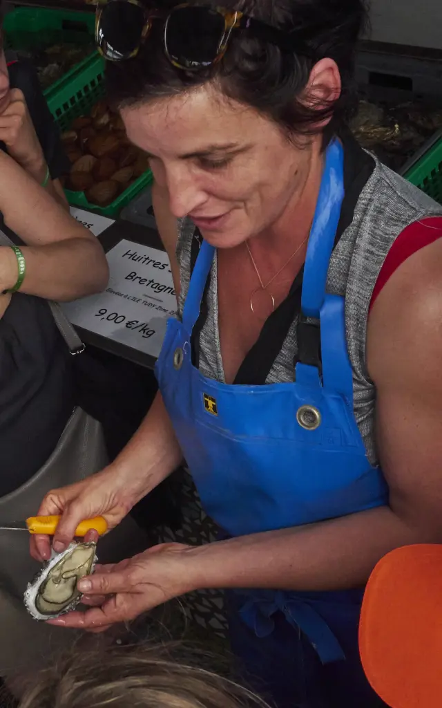 Eén foto toont een groep mensen rond een vrouw met een blauw schort, die een schelp/oestertje in haar handen houdt, alsof ze een demonstratie geeft. Verschillende mensen kijken toe, waaronder iemand op de voorgrond met een witte hoed; in het midden lacht een vrouw met een bril. De afbeelding is ingekaderd in vogelvluchtperspectief, in een omgeving die lijkt op een werk-/verkoopruimte, met groene kratten op de achtergrond.