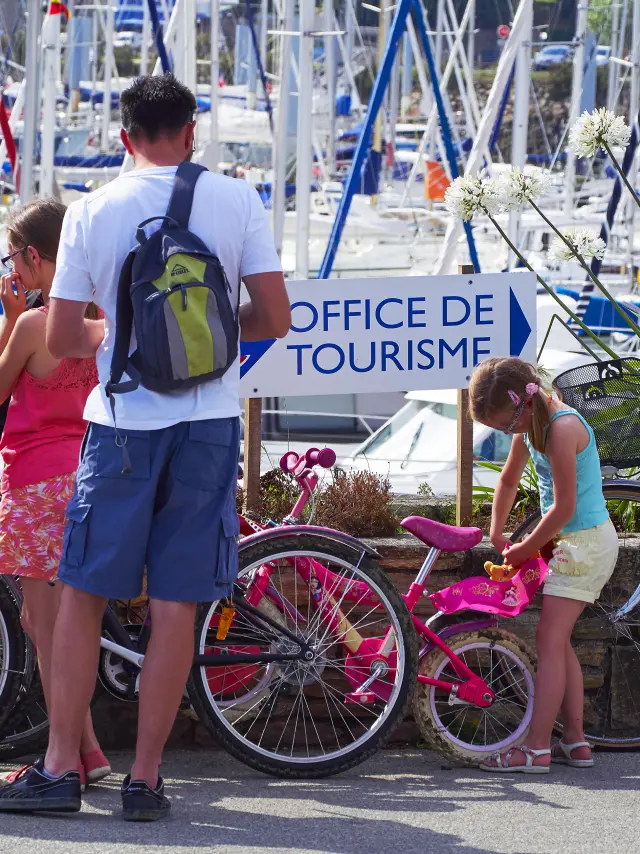 This photo shows a scene in a marina full of boat masts. In the foreground are four people and several bicycles. A woman with brown hair, wearing a red top and blue shorts, is standing next to a bicycle (grey rucksack with a yellow section). A man in a white t-shirt and blue shorts, with a green and grey rucksack, is standing from behind. Another person is sitting or crouching next to the bikes. A child wearing a blue top and light-coloured shorts is leaning over a small pink bicycle. A sign in the middle of the image reads OFFICE DE TOURISME.