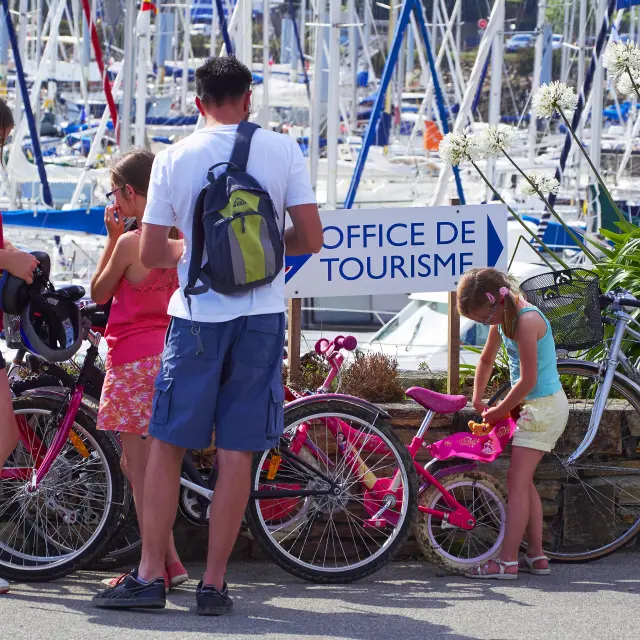 Une photo qui montre une scène dans un port de plaisance rempli de mâts de bateaux. Au premier plan, quatre personnes et plusieurs vélos. Une femme aux cheveux bruns, portant un haut rouge et un short bleu, se tient près d’un vélo (sac à dos gris avec une partie jaune). Un homme en t-shirt blanc et short bleu, avec un sac à dos vert et gris, est debout de dos. Une autre personne est assise ou accroupie près des vélos. Une enfant portant un haut bleu et un short clair est penchée sur un petit vélo rose. Un panneau au milieu de l’image affiche le texte “OFFICE DE TOURISME”.