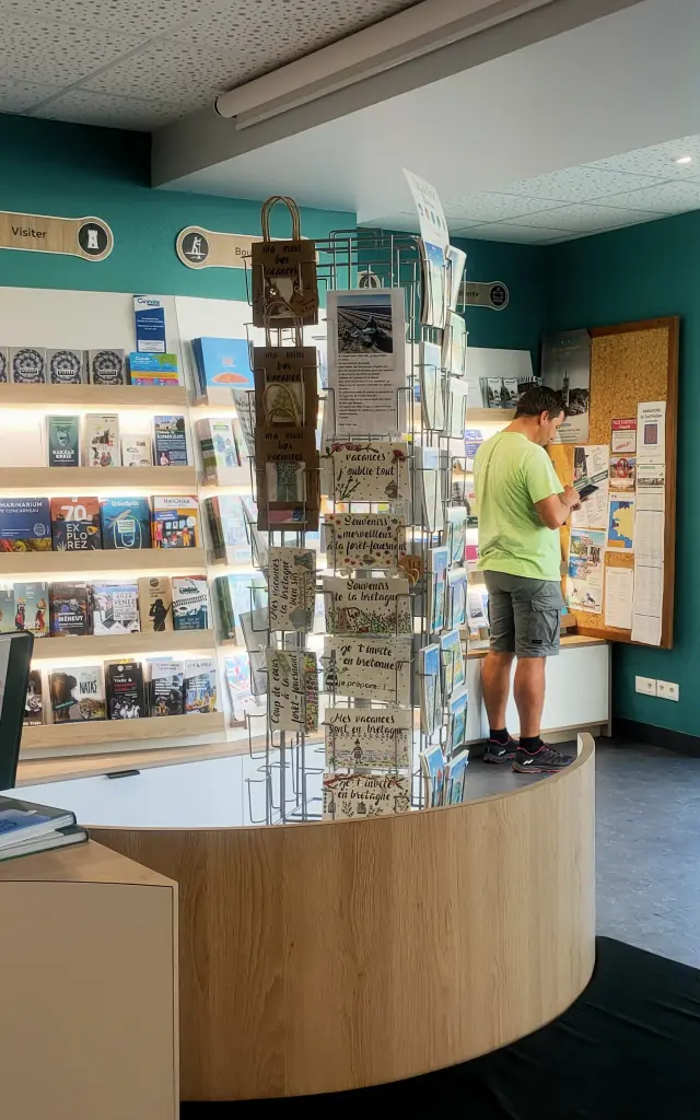 Een foto van het interieur van een VVV-kantoor. De kamer heeft turquoise muren, een gebogen toonbank van licht hout, een display met ansichtkaarten in het midden, planken vol brochures en boeken en een tv-scherm aan de muur. Bij de glazen ingang aan de rechterkant staan twee grote turquoise cilindervormige pilaren. Een persoon staat bij een prikbord aan de achterkant; een ander silhouet is zichtbaar in de richting van de deur.