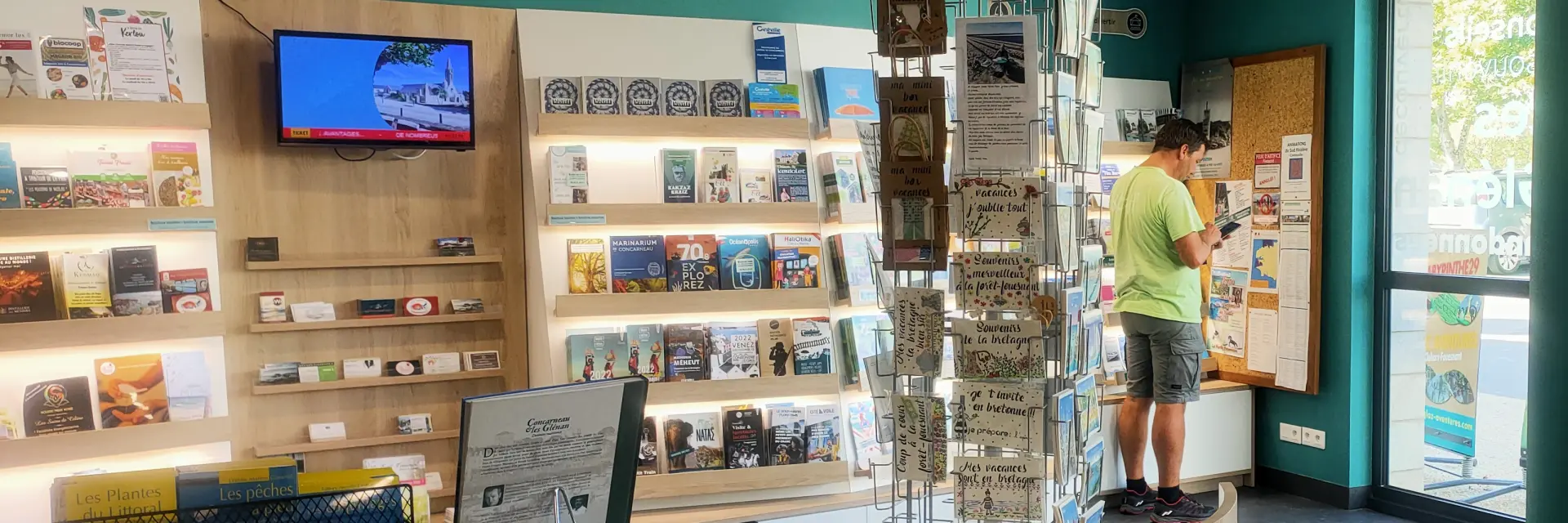 Un photo montrant l’intérieur d’un office de tourisme. La pièce a des murs turquoise, un comptoir en bois clair en forme courbe, un présentoir à cartes postales au milieu, des étagères remplies de brochures et livres, et un écran TV accroché au mur. Deux grands piliers cylindriques turquoise se trouvent près de l’entrée vitrée à droite. Une personne est debout près d’un panneau d’affichage au fond; une autre silhouette est visible vers la porte.