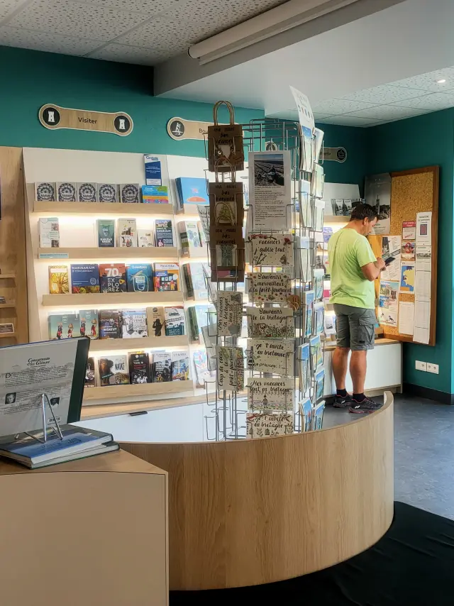 Un photo montrant l’intérieur d’un office de tourisme. La pièce a des murs turquoise, un comptoir en bois clair en forme courbe, un présentoir à cartes postales au milieu, des étagères remplies de brochures et livres, et un écran TV accroché au mur. Deux grands piliers cylindriques turquoise se trouvent près de l’entrée vitrée à droite. Une personne est debout près d’un panneau d’affichage au fond; une autre silhouette est visible vers la porte.